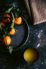 Вид зверху на кухоль зі свіжим фруктовим соком і тарілкою з стиглими мандаринами, розміщеними на грубому чорному столі біля серветки — стокове фото
