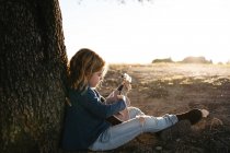 Side view of adorable serious little girl in casual wear playing ukulele guitar while sitting near tree in sunny summer day in countryside — Stock Photo
