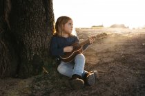 Очаровательная серьезная девочка в повседневной одежде играет на гитаре укулеле, сидя рядом с деревом в солнечный летний день в сельской местности — стоковое фото