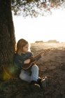 Adorável menina séria em uso casual tocando ukulele guitarra enquanto sentado perto da árvore no dia ensolarado de verão no campo — Fotografia de Stock