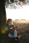 Adorável menina séria em uso casual tocando ukulele guitarra enquanto sentado perto da árvore no dia ensolarado de verão no campo — Fotografia de Stock