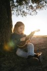 Adorable serious little girl in casual wear playing ukulele guitar while sitting near tree in sunny summer day in countryside — Stock Photo