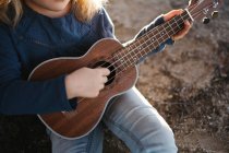 Обрізана невпізнавана маленька дівчинка в повсякденному одязі грає на укулелевій гітарі, сидячи біля дерева в сонячний літній день у сільській місцевості — стокове фото