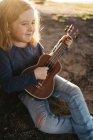 Adorable niña seria en ropa casual tocando la guitarra ukelele mientras está sentado cerca del árbol en el soleado día de verano en el campo - foto de stock