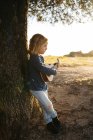 Adorable serious little girl in casual wear playing ukulele guitar while standing near tree in sunny summer day in countryside — Stock Photo
