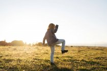 Visão traseira de anônimo adorável menina ativa em casual wear jogando e dançando no campo verde enquanto desfruta de noite de verão ensolarado no campo — Fotografia de Stock