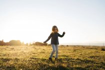 Очаровательная активная девочка в повседневной одежде играет и танцует на зеленом поле, наслаждаясь солнечным летним вечером в сельской местности — стоковое фото
