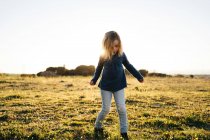 Adorable niña activa en ropa casual jugando y bailando en el campo verde mientras disfruta de la soleada noche de verano en el campo - foto de stock