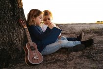 Vista lateral de la adorable niña leyendo interesante historia a su hermano menor mientras se sientan juntos bajo el árbol con la guitarra ukelele en el día de verano en el campo - foto de stock