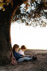 Вид сбоку восхитительной маленькой девочки, читающей интересную историю младшему брату, сидя вместе под деревом с гитарой в летний день в деревне — стоковое фото