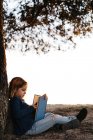 Вид збоку спокійна маленька дівчинка в повсякденному одязі читає казкову книгу, сидячи під деревом в полі в сонячний літній вечір — стокове фото