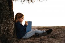 Side view of calm little girl in casual wear reading fairy tale book while sitting under tree in field in sunny summer evening — Stock Photo