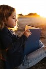 Вид сбоку спокойной маленькой девочки в повседневной одежде, читающей сказку, сидя под деревом в поле солнечным летним вечером — стоковое фото