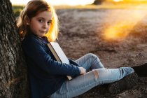 Вид сбоку спокойной маленькой девочки в повседневной одежде, смотрящей на камеру, читающей сказку, сидя под деревом в поле солнечным летним вечером — стоковое фото