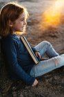Вид збоку спокійна маленька дівчинка в повсякденному одязі дивиться в очі посміхаючись тримає казкову книгу, сидячи під деревом на полі в сонячний літній вечір — стокове фото