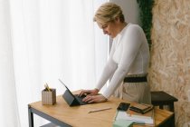 Mujer adulta rubia enfocada de pie en la mesa de madera con papelería escribiendo en el teclado portátil de la tableta contra la ventana de luz - foto de stock