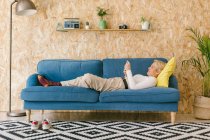Вид сбоку на стильную блондинку-предпринимательницу, лежащую на голубом диване в современном уютном офисе с телефоном в перерыве — стоковое фото