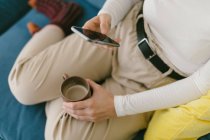Faceless copped woman without shoes chilling on cozy sofa in office enjoying cup of coffee and surfing mobile phone — Stock Photo