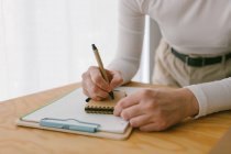 Безликая деловая женщина, нагибающаяся на деревянный стол и пишущая в блокноте ручкой — стоковое фото