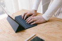 Обрізані руки невпізнаваної людини за дерев'яним столом за допомогою планшета з клавіатурою, що працює в спокійному затишному офісі — стокове фото