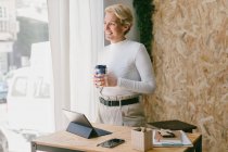 Посміхаючись щаслива доросла жінка за робочим столом насолоджується ранковою кавою, посміхаючись у вікні офісу — стокове фото