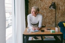 Сосредоточенная взрослая деловая женщина отворачивается, делая заметки о плане на планшете, нагибаясь на деревянном столе в светлом офисе — стоковое фото