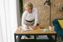 Сосредоточенная взрослая деловая женщина, записывающая план в буфер обмена, нагибающаяся на деревянный стол в светлом кабинете — стоковое фото
