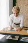 Focused adult businesswoman taking notes of plan on clipboard bending on wooden table in light office — Stock Photo