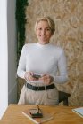 Happy focused adult businesswoman looking at camera while using mobile phone near notepad on wooden table in light office — Stock Photo