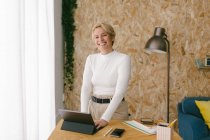 Lässige moderne blonde Geschäftsfrau lächelt in die Kamera am Holztisch mit Tablet und Tastatur, die in einem ruhigen, gemütlichen Büro arbeitet — Stockfoto