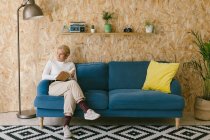 Blonde woman with short hair in white shirt sitting on sofa looking away and writing in notebook working on business project — Stock Photo