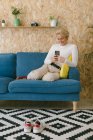 Vista lateral de la mujer alegre informal mensajes de texto en el teléfono inteligente mientras tiene descanso en la oficina sentado en el sofá - foto de stock