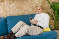 Вид сбоку случайной женщины, текстурирующей на смартфоне во время перерыва в работе сидя на диване — стоковое фото
