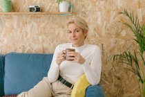Ruhige erwachsene Geschäftsfrau mit kurzen blonden Haaren sitzt auf dem gemütlichen Sofa im Büro, trinkt Kaffee und lächelt gelassen in die Kamera — Stockfoto
