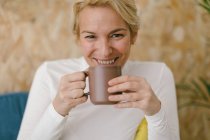 Спокойная взрослая деловая женщина с короткими светлыми волосами, сидящая на уютном диване в офисе, выпивая кружку кофе и спокойно улыбаясь в камеру — стоковое фото