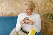 Calma donna d'affari adulta con capelli corti biondi seduta su un accogliente divano in ufficio con tazza di caffè e sorridente con calma guardando altrove — Foto stock