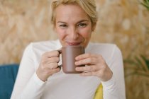 Femme d'affaires adulte calme avec des cheveux blonds courts assis sur un canapé confortable dans le bureau ayant tasse de café et souriant calmement à la caméra — Photo de stock