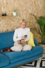 Блондинка-бизнесвумен с короткими волосами без обуви, отдыхающая на уютном диване в офисе, наслаждаясь чашечкой кофе и серфингом на мобильном телефоне — стоковое фото