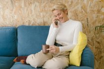 Блондинка-бизнесвумен с короткими волосами без обуви, отдыхающая на уютном диване в офисе, наслаждаясь чашечкой кофе и серфингом на мобильном телефоне — стоковое фото