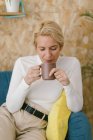 Mulher de negócios adulta calma com cabelo loiro curto sentado no sofá acolhedor no escritório tendo caneca de café e sorrindo calmamente olhando para longe — Fotografia de Stock