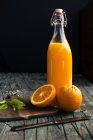 Garrafa de suco de laranja cítrico fresco colocada perto de metades de laranjas frescas em uma mesa rústica escura de madeira no fundo escuro — Fotografia de Stock