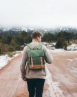 Back view of young male traveler in sweater with backpack walking on dirt rural road in cloudy winter day with green forest and snow on roadsides — Stock Photo