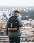 Вид сзади на молодого туриста в теплой верхней одежде с рюкзаком, стоящим на обочине проселочной дороги со снежным полем и лесом на заднем плане в пасмурный день — стоковое фото
