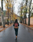Обратный вид молодой активной женщины в повседневной одежде с рюкзаком, идущей по пустой асфальтовой дороге в осеннем парке в пасмурный день — стоковое фото