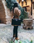 Vista lateral de jovem viajante do sexo feminino em roupa elegante com saco no ombro em pé no quintal do edifício de pedra velha no dia de outono na cidade — Fotografia de Stock