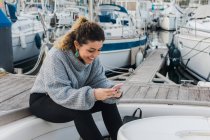 Vue latérale de la jeune femme en pull décontracté souriant tout en surfant sur téléphone mobile dans un yacht moderne — Photo de stock