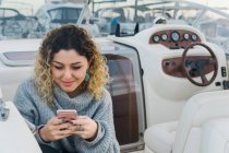 From above of young woman in casual sweater smiling while surfing on mobile phone in modern yacht — Stock Photo