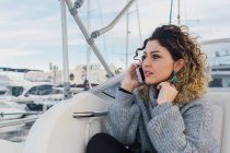 Jeune femme en pull décontracté souriant tout en surfant sur un téléphone mobile dans un yacht moderne — Photo de stock