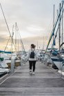 Вид ззаду на безлику молоду жінку в повсякденному одязі з чорним рюкзаком, що йде на дерев'яному пірсі між сучасними яхтами на заході сонця — стокове фото