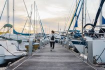 Vista posteriore di giovane donna senza volto in abbigliamento casual con zaino nero che cammina su un molo di legno tra yacht moderni al tramonto — Foto stock